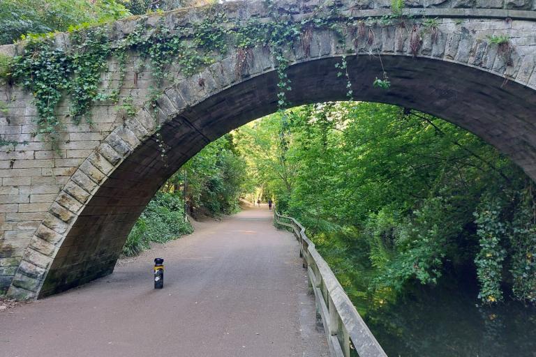 Photo of a wide path alongside a river going under a stony bridge.