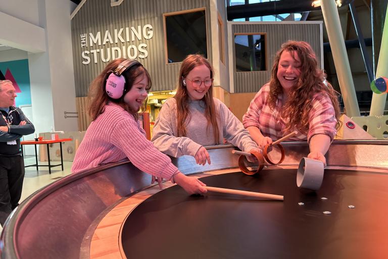 Two smiling girls, one of them with ear defenders, and a women hold wooden sticks on a black round surface. Behind them is a sign reading The Making Studios