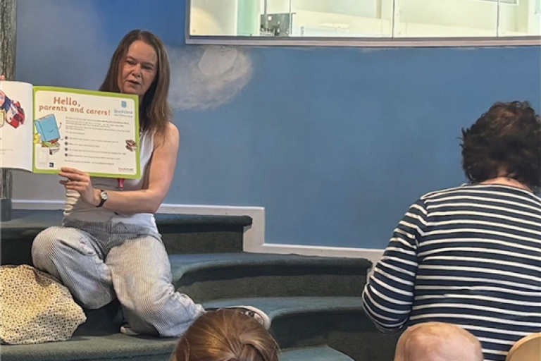 Photo of a woman holding a book in front of some children and adults at Newcastle City Library. 