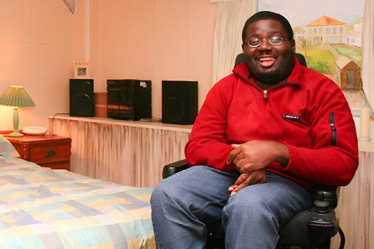 A man in a red jacket sitting in a motorized wheelchair in a cozy bedroom.