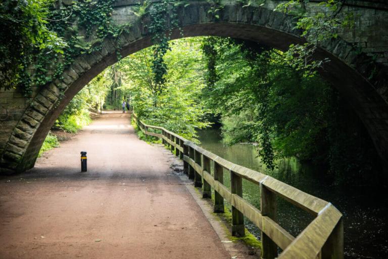 Photo of a wide path alongside a river going under a stony bridge.