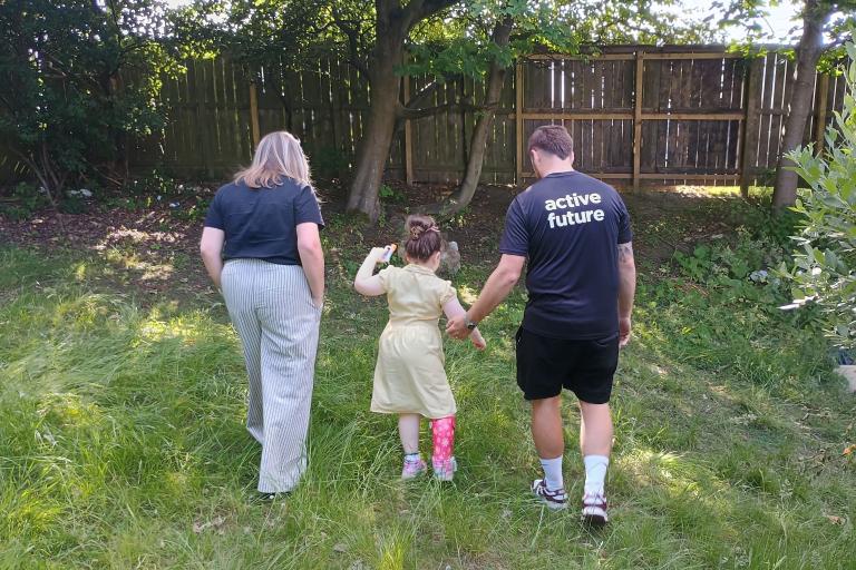 two adults and child walking across the grass towards a tree and a fence. 