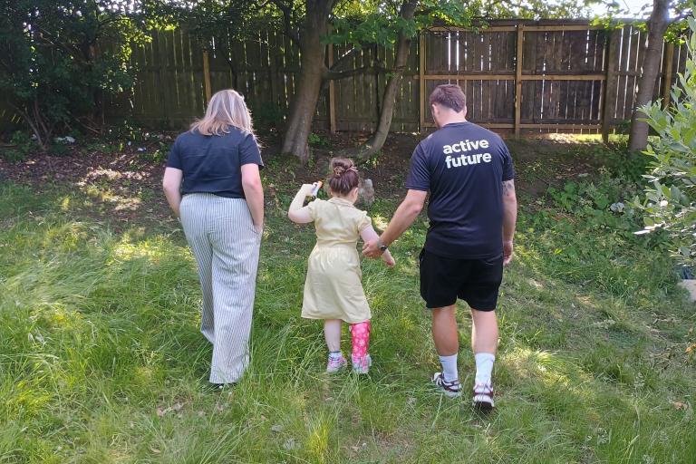two adults and child walking across the grass towards a tree and a fence. 