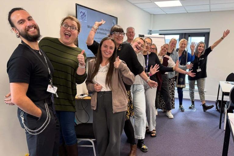A line of adults standing in a classroom setting smiling and raising their arms for the photo.
