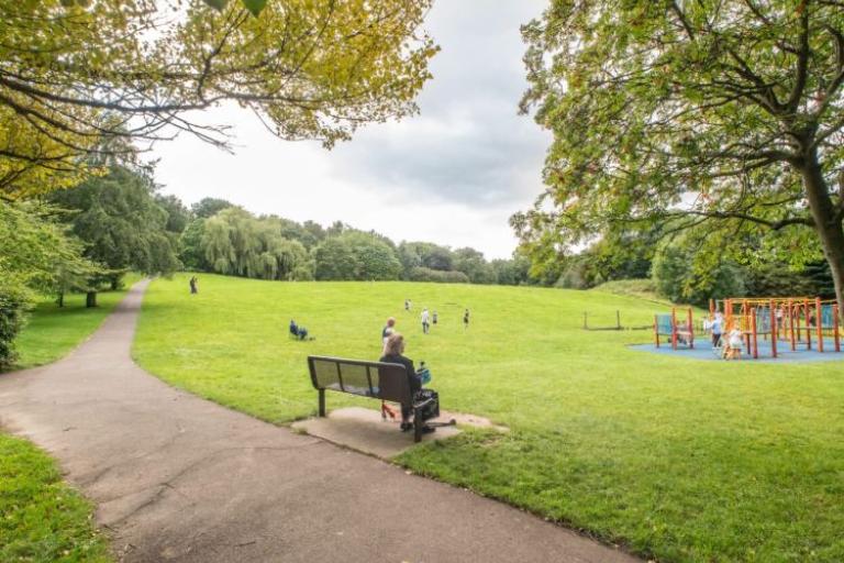 Image of Heaton Park. Person sitting on a bench looking over a children's play area. 