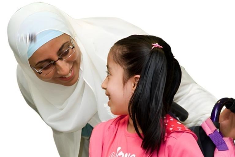 Woman in headscarf smiling at a girl in a wheelchair wearing a pink jacket.