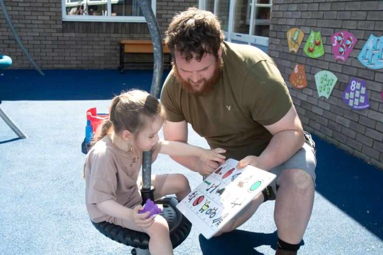 A man holding a communication board as a girl points at one of the pictures on it.