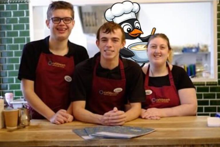 Image includes one of Girtrood's friends. 3 young people smiling and wearing aprons with a cartoon penguin wearing a chef's hat in the background