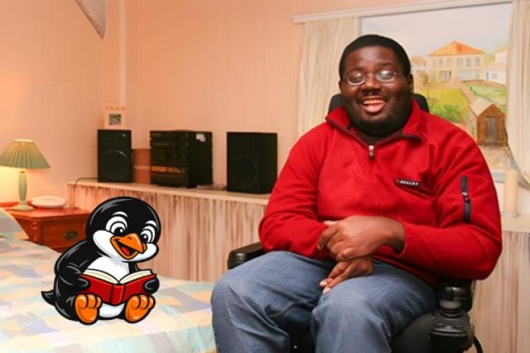 A smiling man in a bedroom sat on an electric wheelchair with a cartoon penguin sat on the bed reading a book