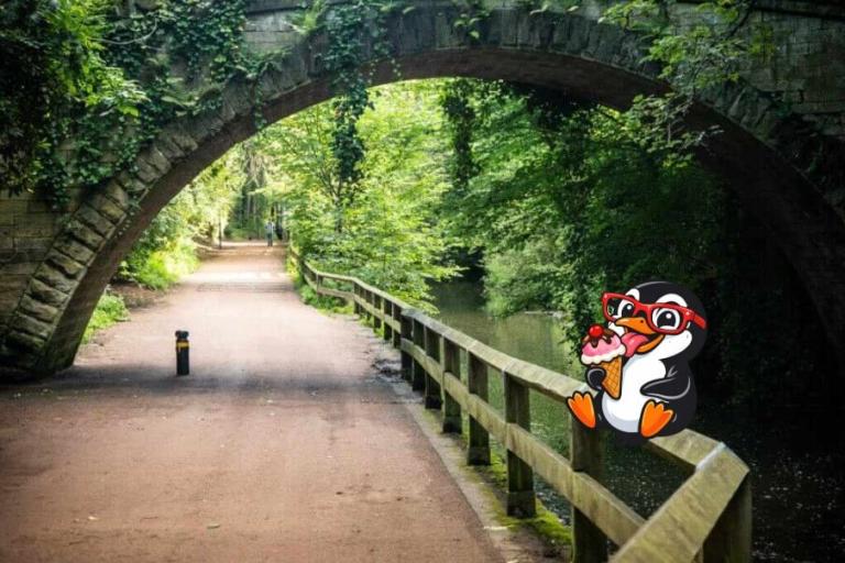 A path going under a stone bridge on a sunny day. A cartoon penguin is eating an ice cream on the fence beside the path. 