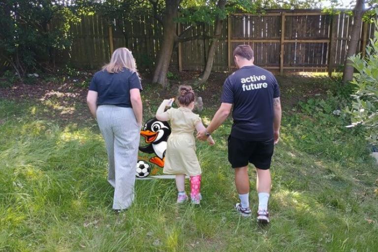 A man, woman and child walking away across a grassy field. A cartoon penguin kicking a football can be seen behind these people.