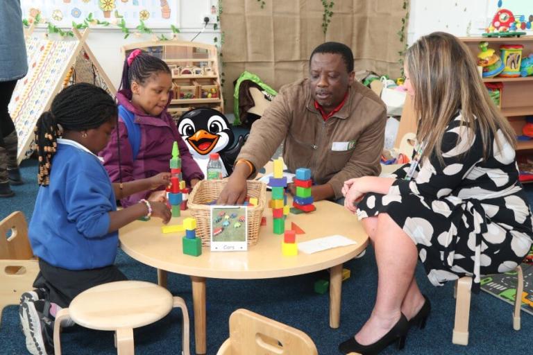 A father and children playing with blocks at a table as a woman watches. A cartoon penguin is waving from behind the table. 