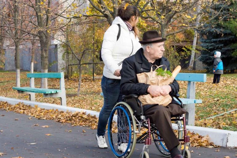 Lady pushing disabled elderly man. Lady is looking at young child in background 