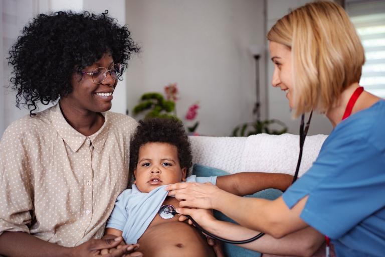 Nurse with stethoscope examines baby who is sat on carers knee