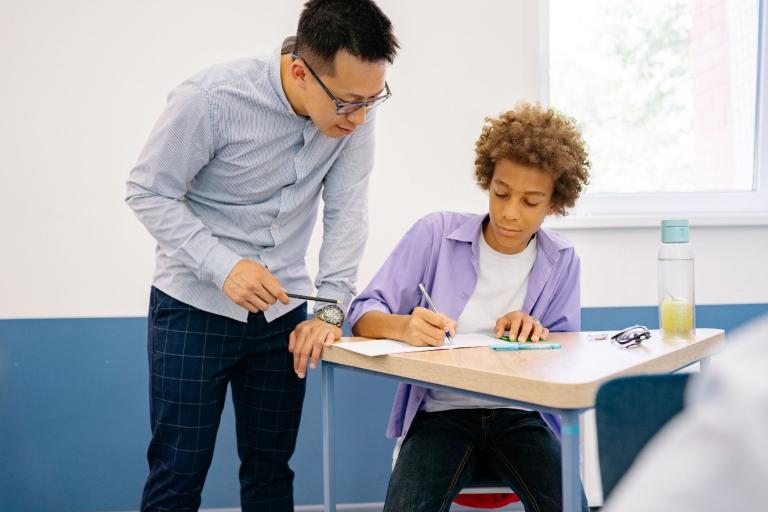 Teenage boy working at desk. Teacher standing next to him checking his work