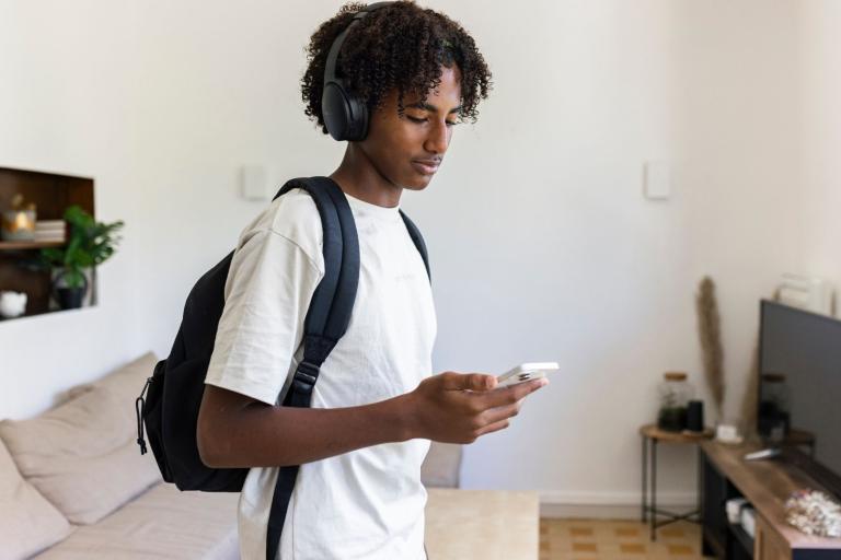 Young man standing in living room holding phone with rucksack over his shoulder