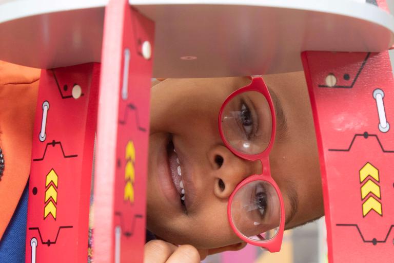 A child wearing glasses looking through a wooden toy space rocket