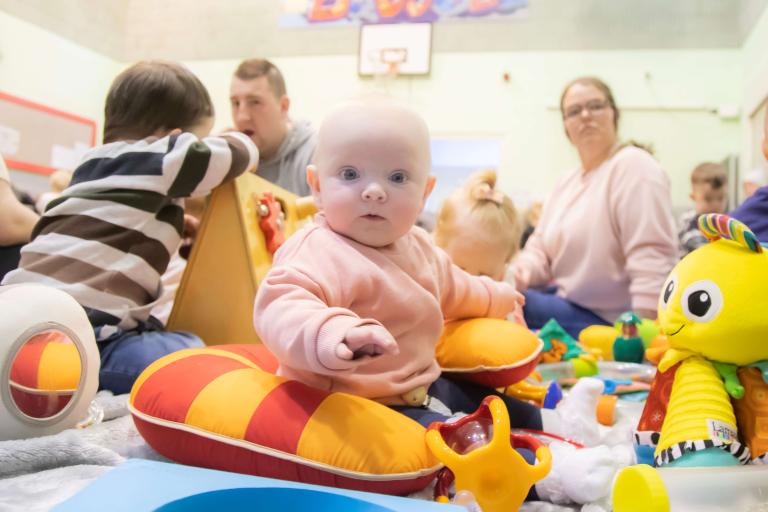 baby sitting in seat cushion the foreground, with family member and toy around