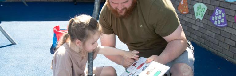 A man holding a communication board as a girl points at one of the pictures on it.