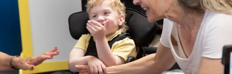 A smiling child in a modified chair holding the arm of a smiling woman.