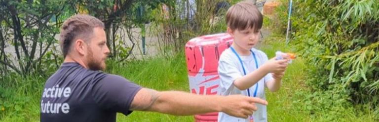 Boy standing outside aiming a nerf gun as an adult points to where he should aim