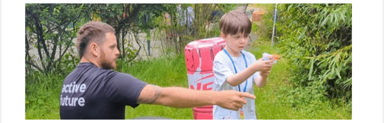 Boy standing outside aiming a nerf gun as an adult points to where he should aim