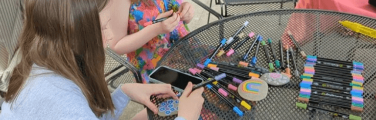 Girls sat at outdoor table with many pens decorating a smooth stone