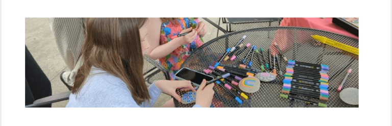 Girls sat at outdoor table with many pens decorating a smooth stone