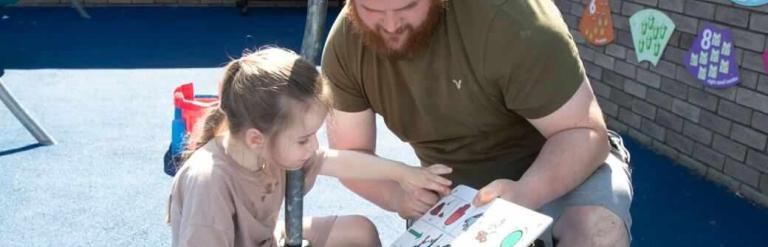 A man holding a communication board as a girl points at one of the pictures on it.